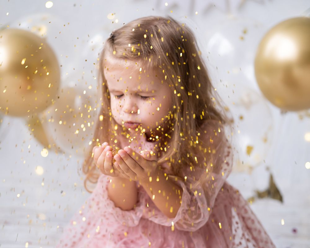 Niña rubia con los ojos cerrados soplando confeti dorado. Al fondo se ven globos dorados.